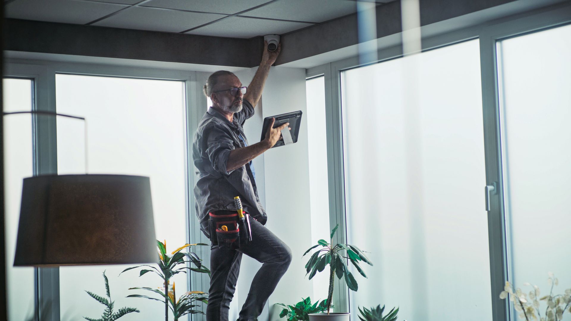 Worker installing a ceiling light in a bright room with large windows