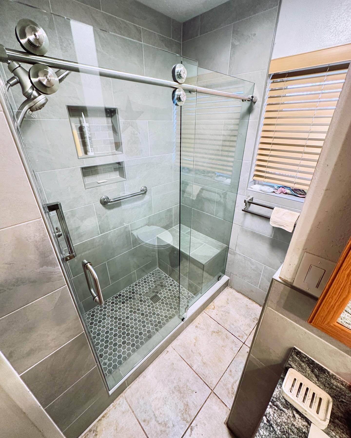 Glass walk-in shower with gray tile walls, chrome fixtures, and a small window blind nearby