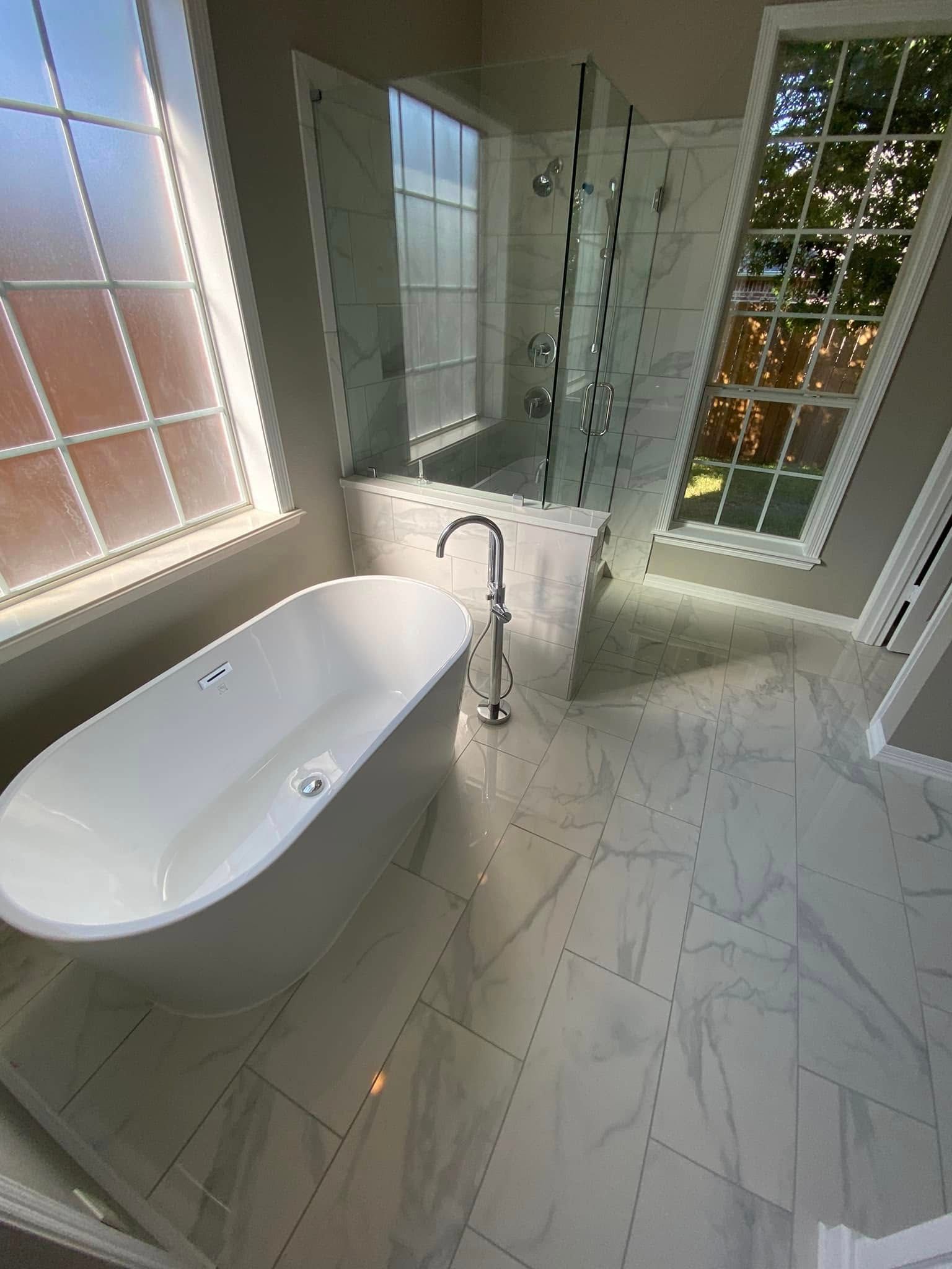 Modern bathroom with a freestanding tub, glass shower, marble tile, and large windows.