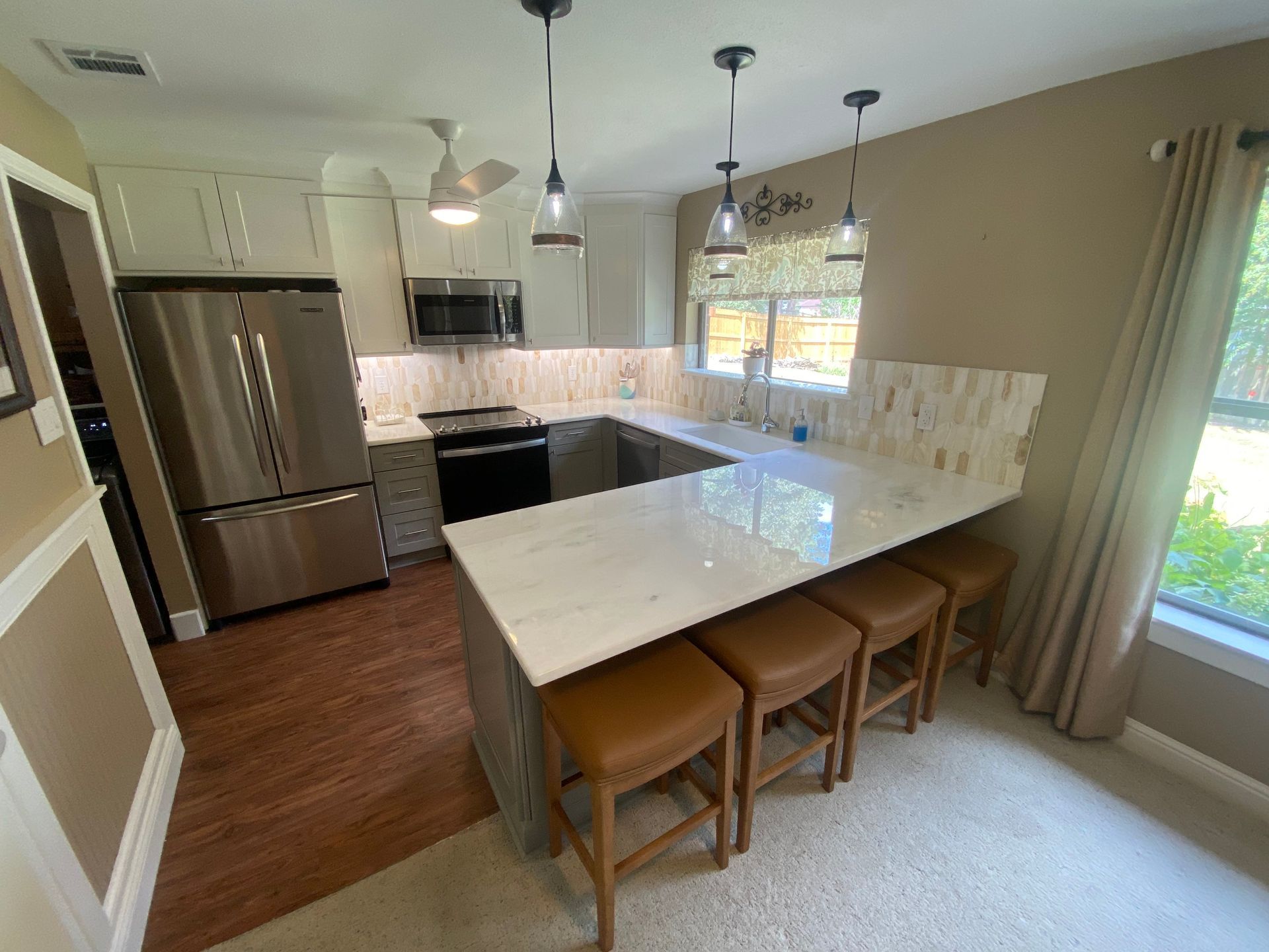 Modern kitchen with white island, pendant lights, stainless fridge, and three wooden stools by a window.