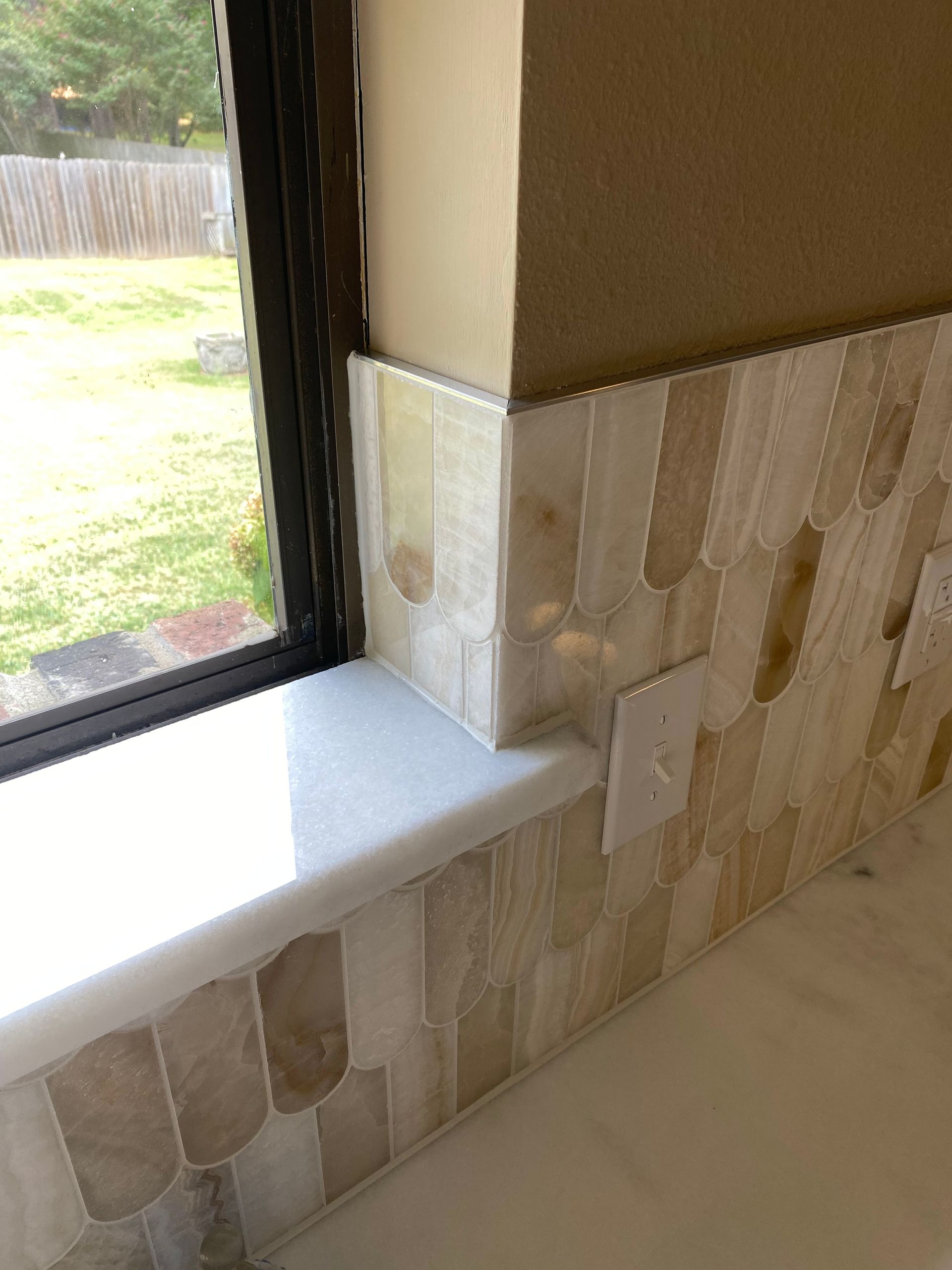 Corner of a tiled window seat with beige stone tiles beside a window and grassy yard outside