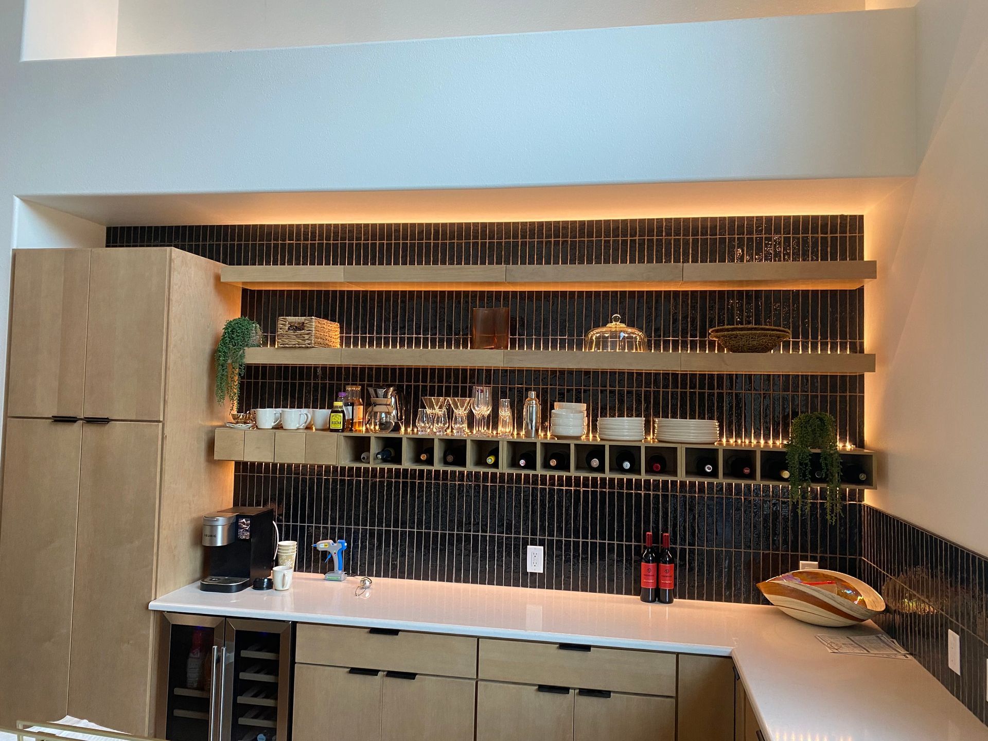 Modern home bar with shelves of bottles, glasses, and decor in a warm-lit alcove.