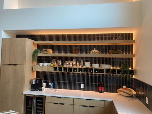 Modern home bar with black shelving, bottles, glassware, and warm lighting against a white wall