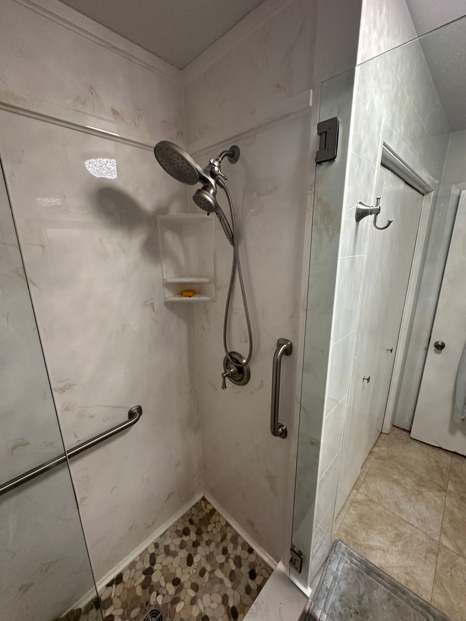 Walk-in shower with handheld showerhead, grab bar, soap shelf, and pebble-tile floor in a white bathroom.