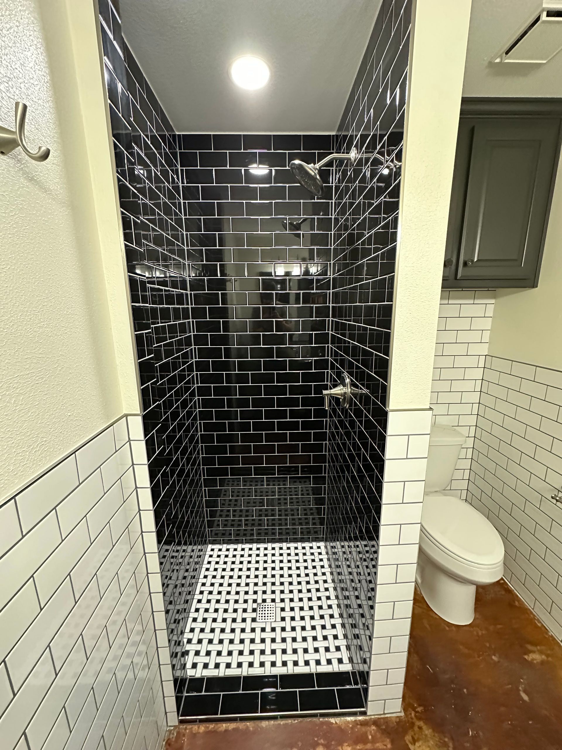 Small tiled shower with black subway walls, white mosaic floor, and a toilet beside it in a bathroom.