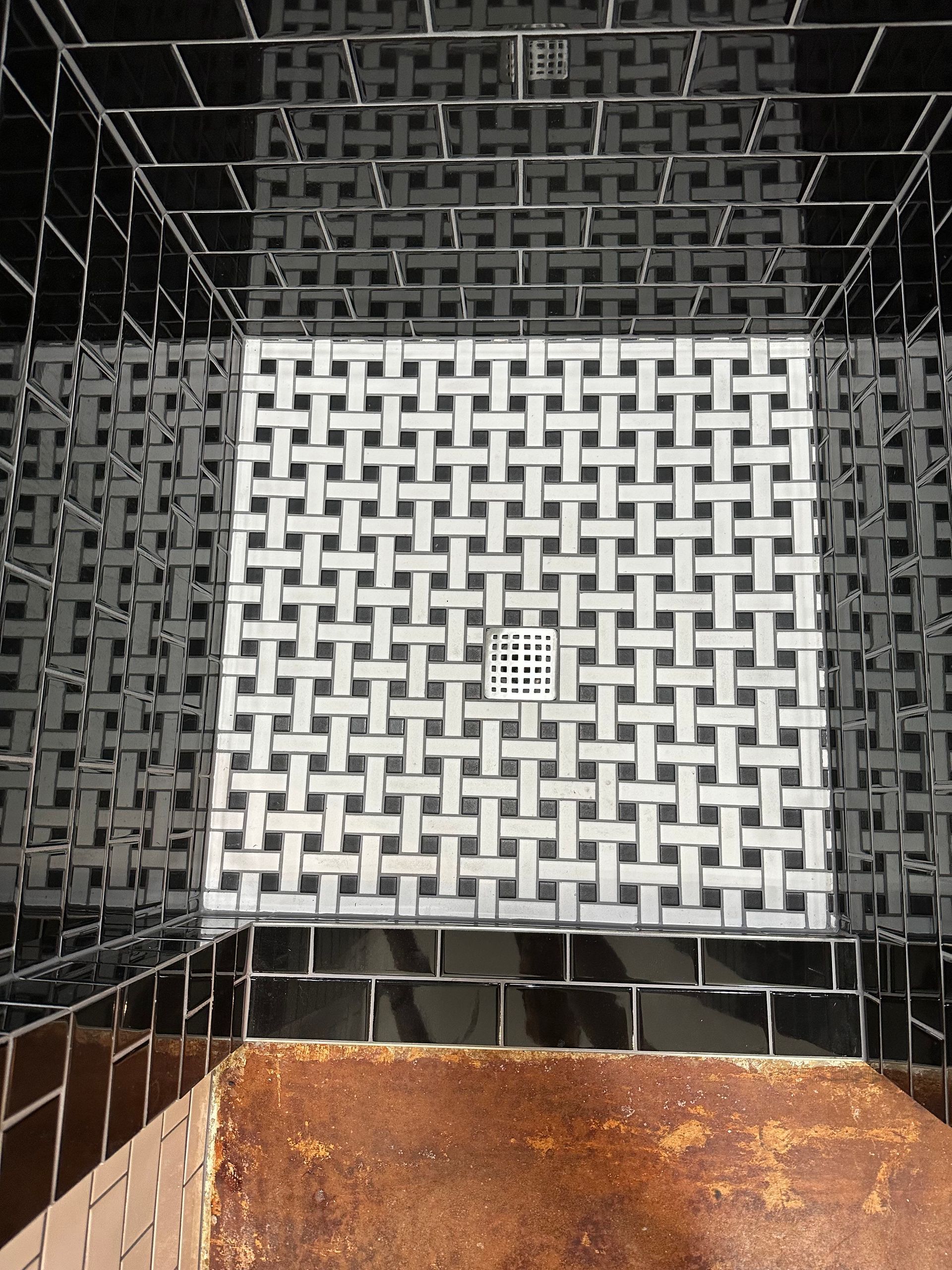 Empty tiled shower corner with black walls, white floor drain cover, and rust-stained base.