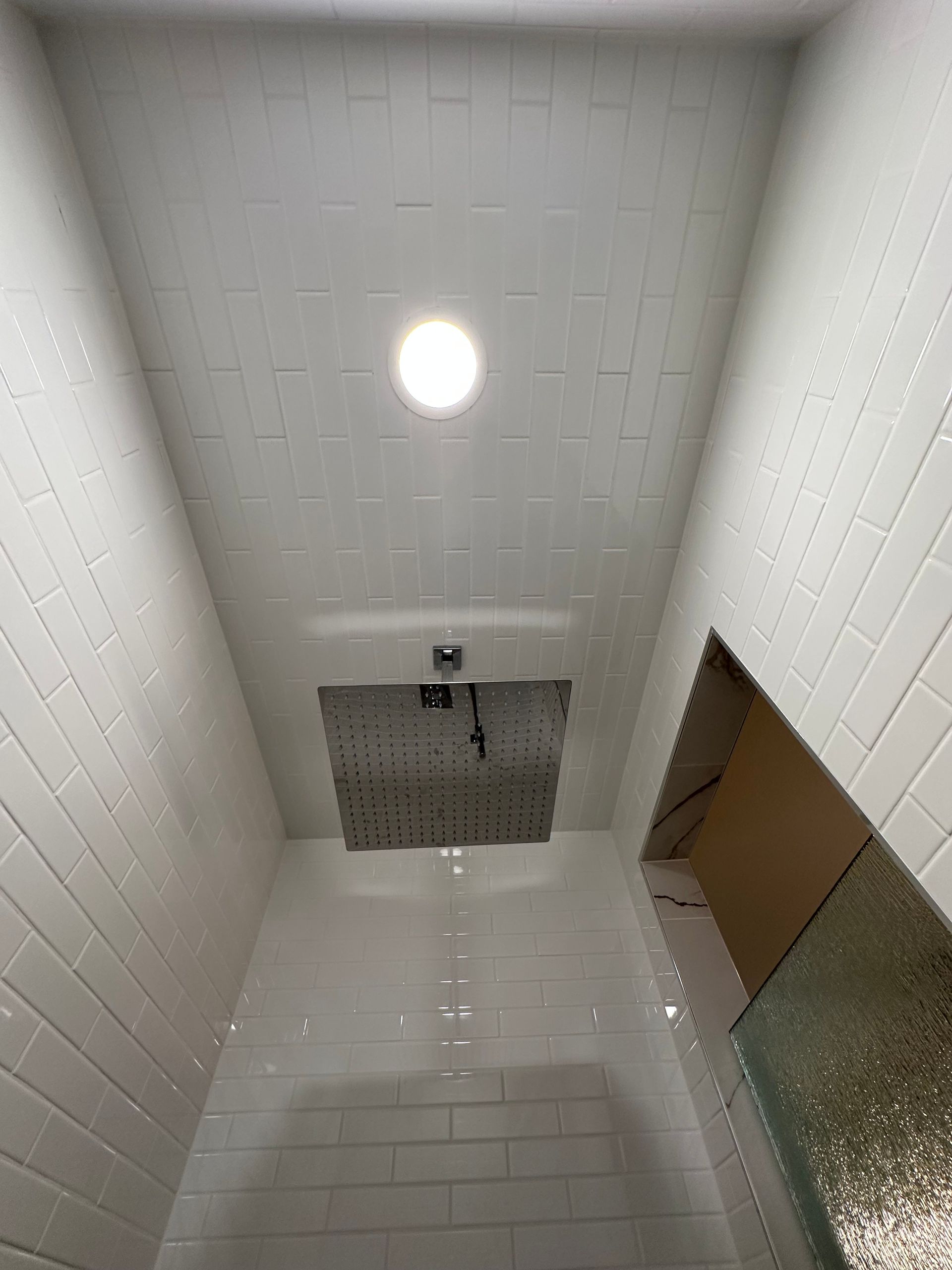 Narrow tiled shower stall with a round ceiling light and a drain on the floor