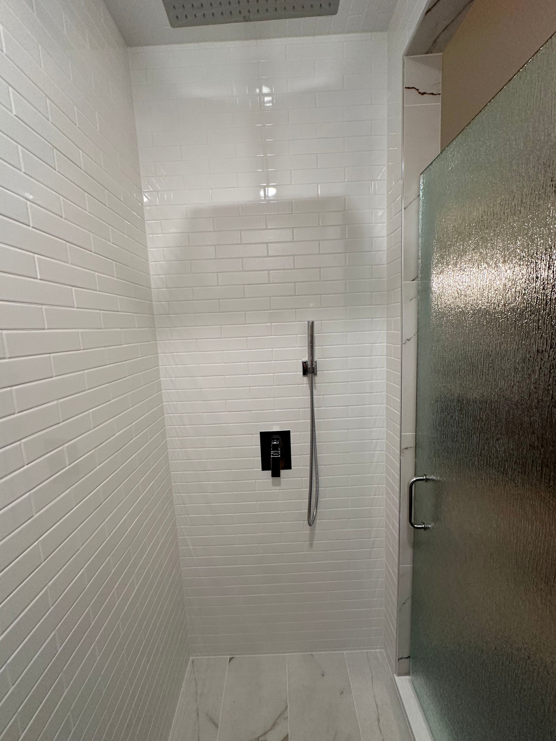 Tiled shower stall with handheld showerhead and frosted glass door open on the right.