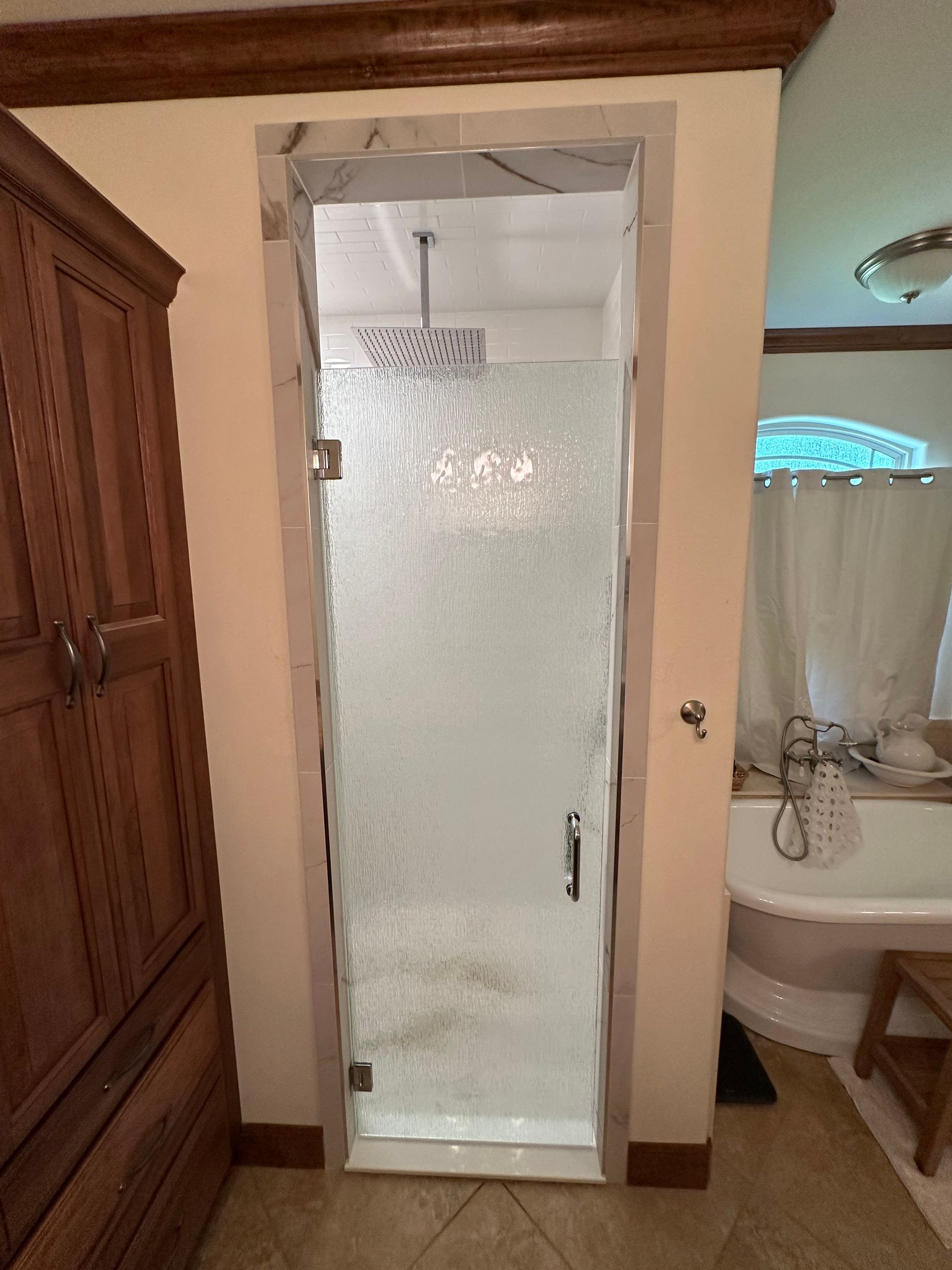 Frameless glass shower stall with tiled walls in a bathroom, beside a sink and wooden cabinet.