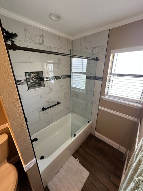 Modern bathroom with glass shower-tub combo, wood floor, and window light