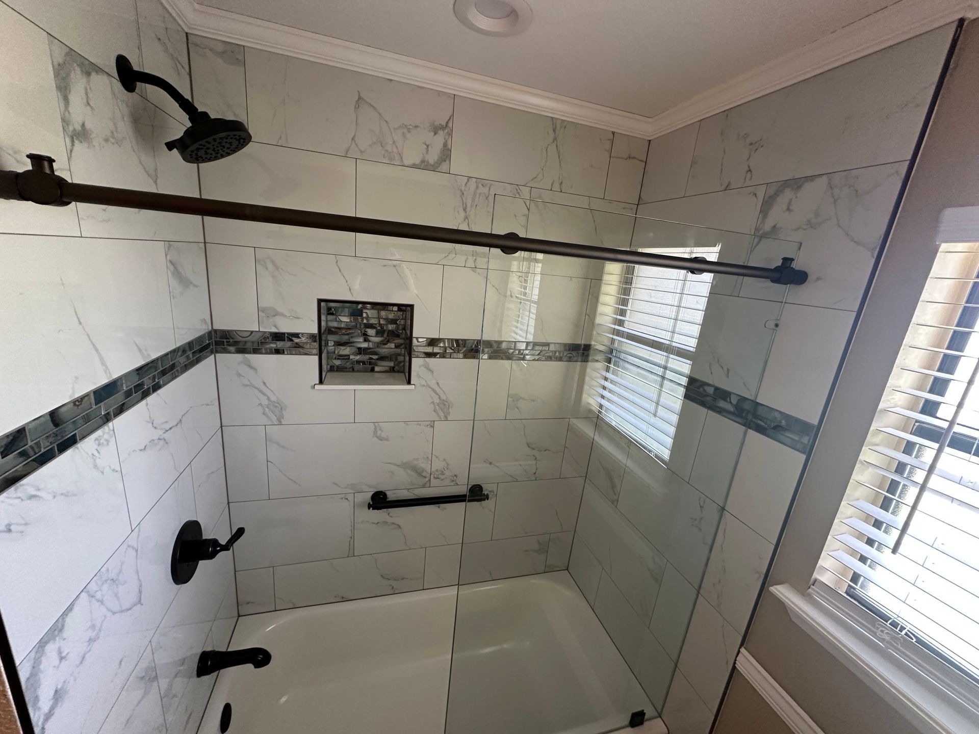 Empty white tiled bathroom shower with bench, grab bars, and window blinds