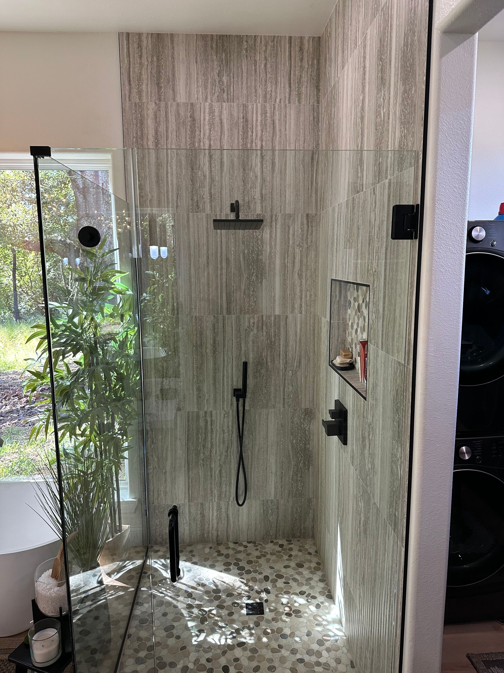 Modern shower with glass wall, gray tile, black fixtures, and pebbled floor beside a window.