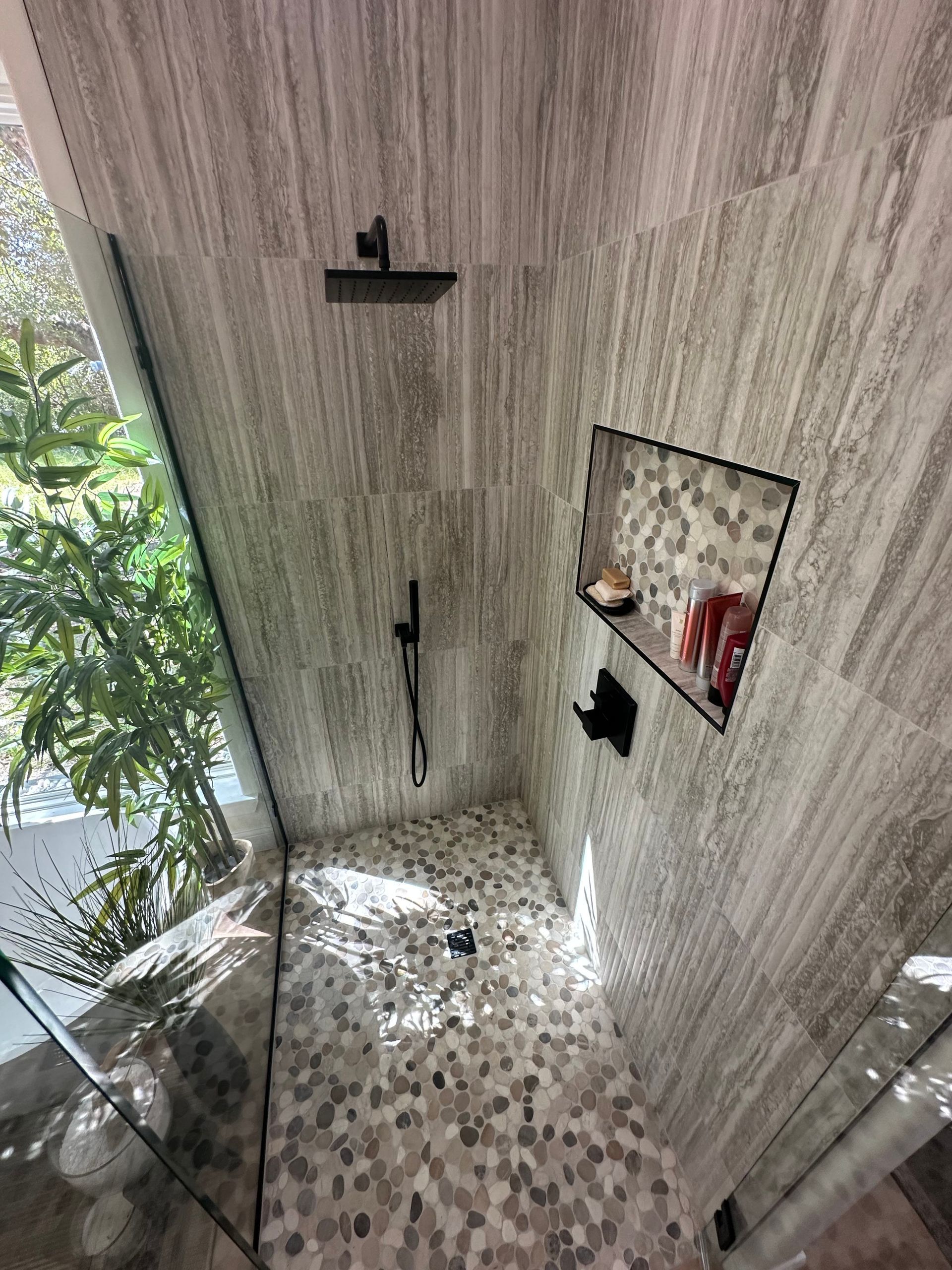 Modern shower with gray tile walls, pebble floor, black fixtures, and a glass door near a window