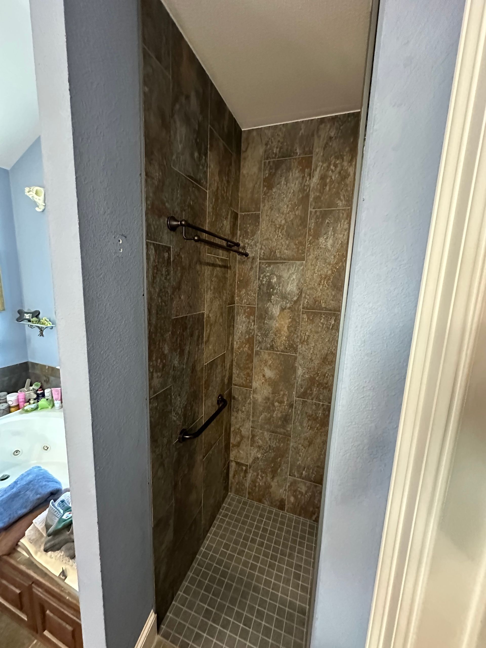 Open shower stall with brown tile walls, gray mosaic floor, and black grab bars