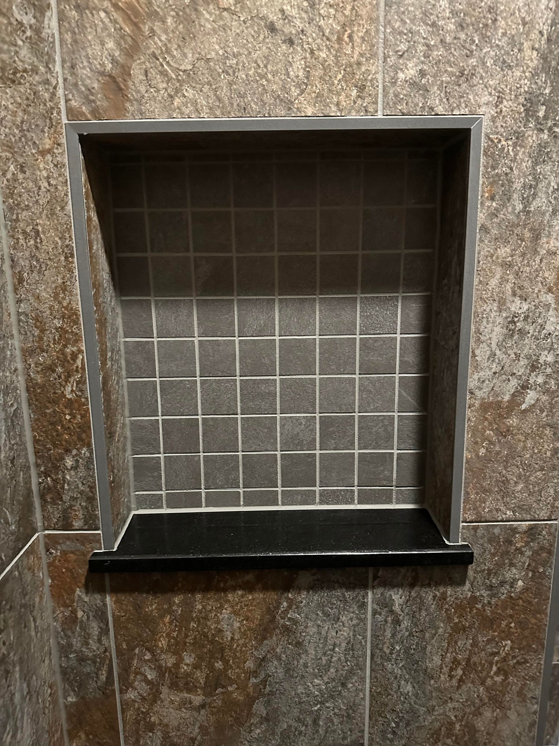 Empty recessed wall niche with gray tile backing and a black shelf against brown stone tile walls