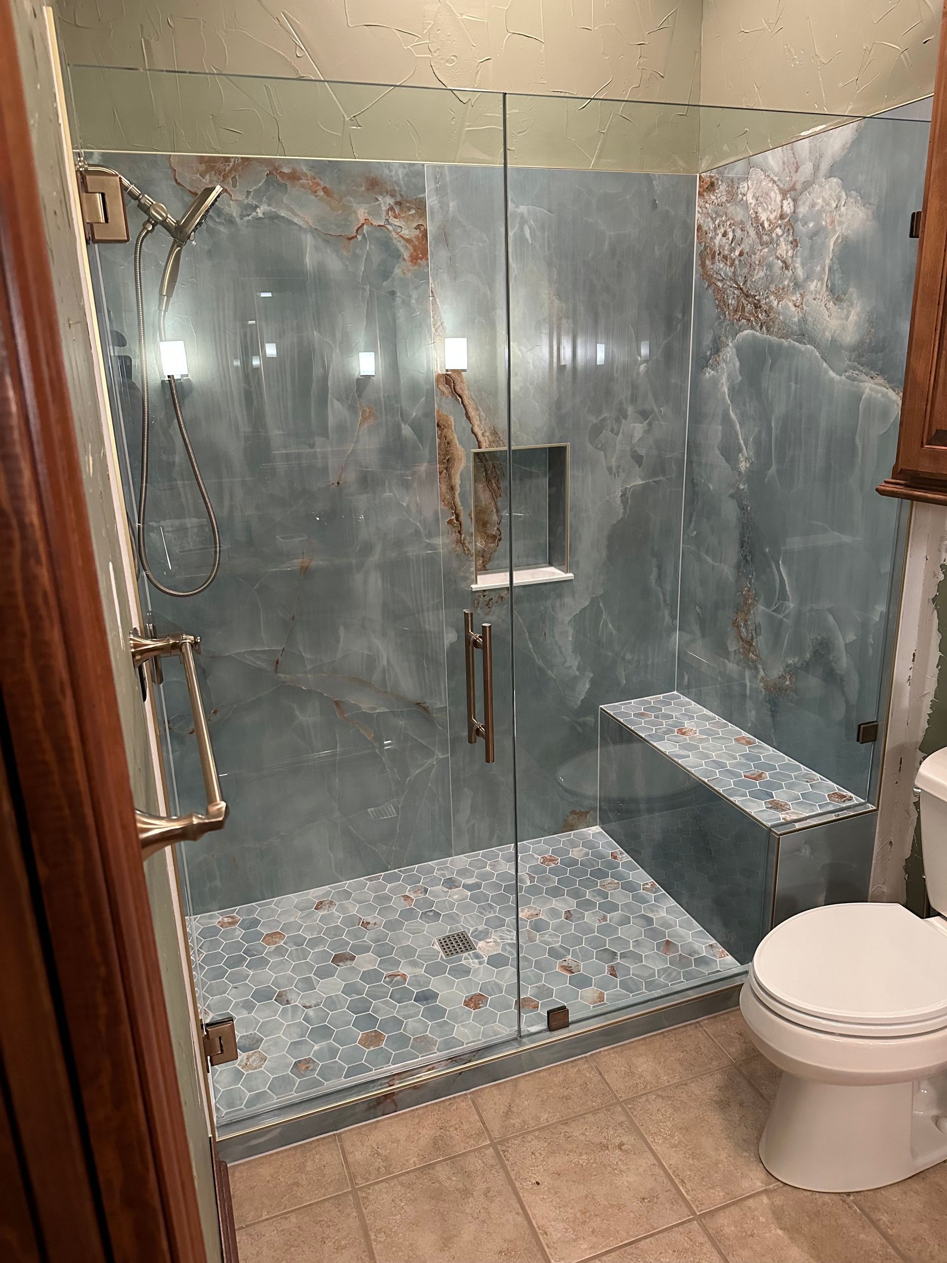 Damaged glass shower stall with blue tile, a built-in bench, and a toilet in a small bathroom.
