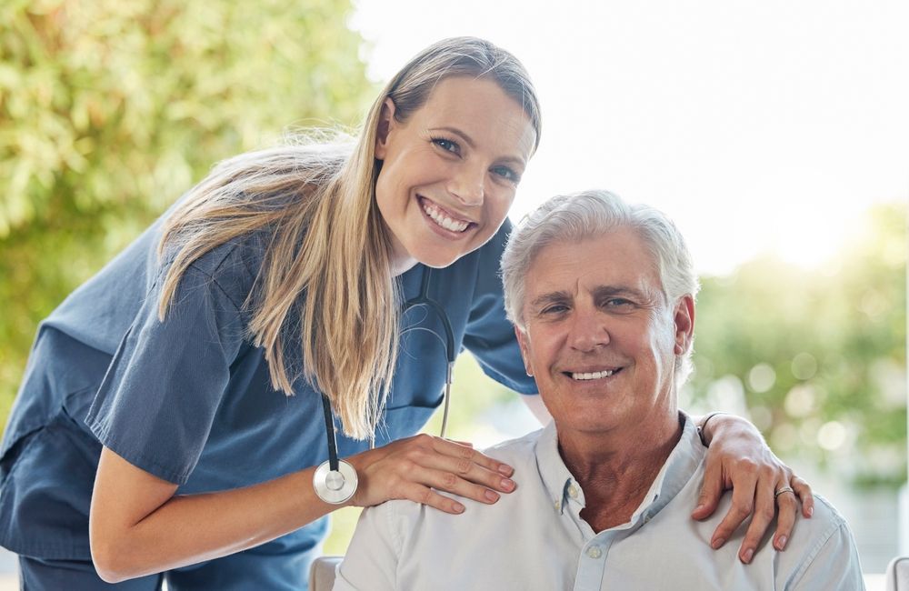 Woman in blue scrubs smiles, arm around a man in a white shirt. Both are smiling outdoors.