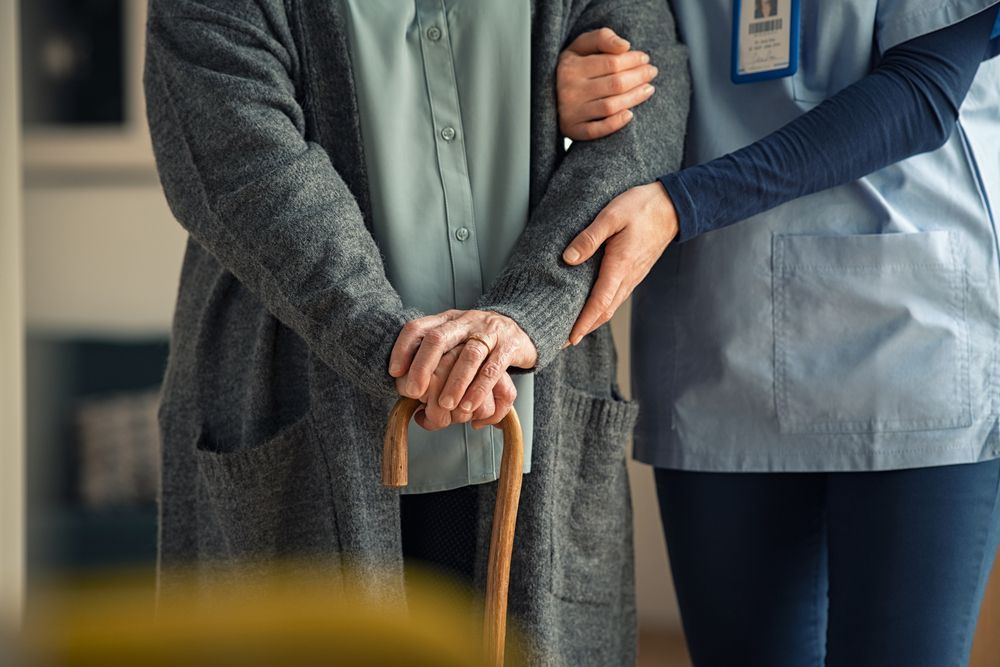 A person using a cane is assisted by a caregiver wearing blue scrubs.
