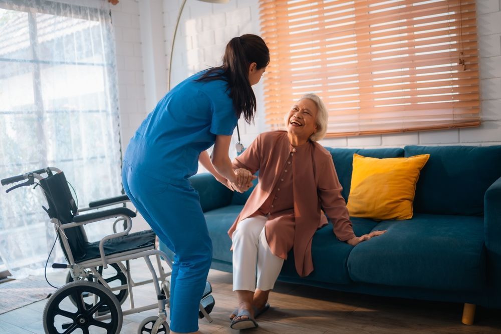 Caregiver assisting an older person to stand up from a couch; wheelchair nearby.