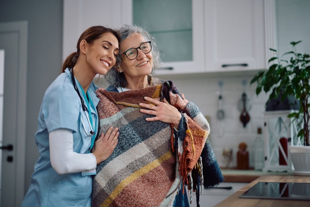 Caregiver embraces older woman wrapped in a blanket in a kitchen, both smiling.