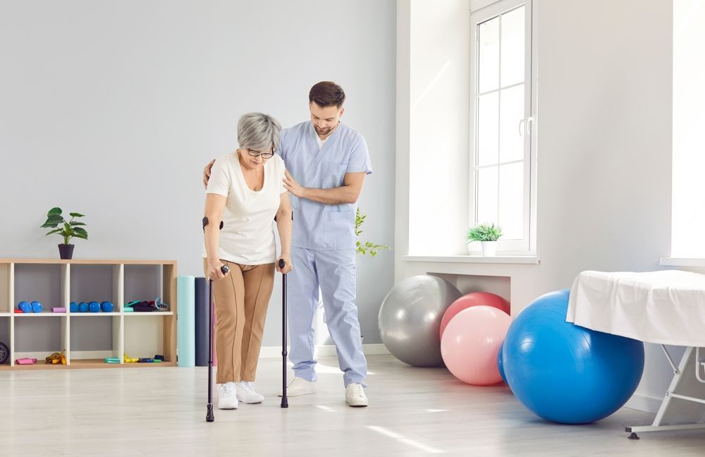 Therapist assisting a person with a cane to walk in a rehabilitation room, blue and gray setting, therapy balls visible.