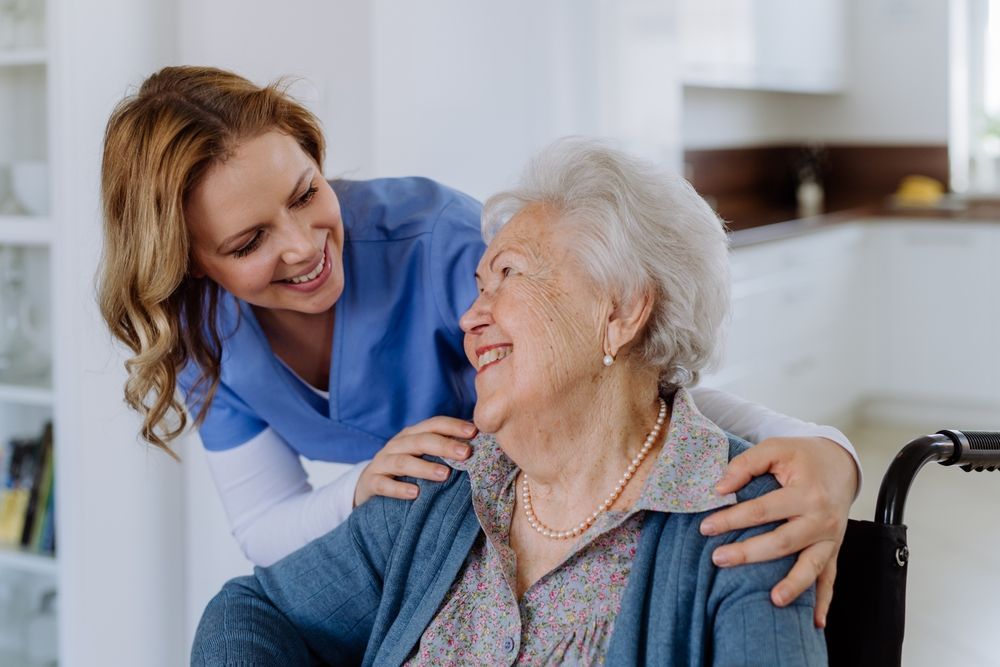 Woman in blue scrubs smiles at older woman in a wheelchair, indoors.
