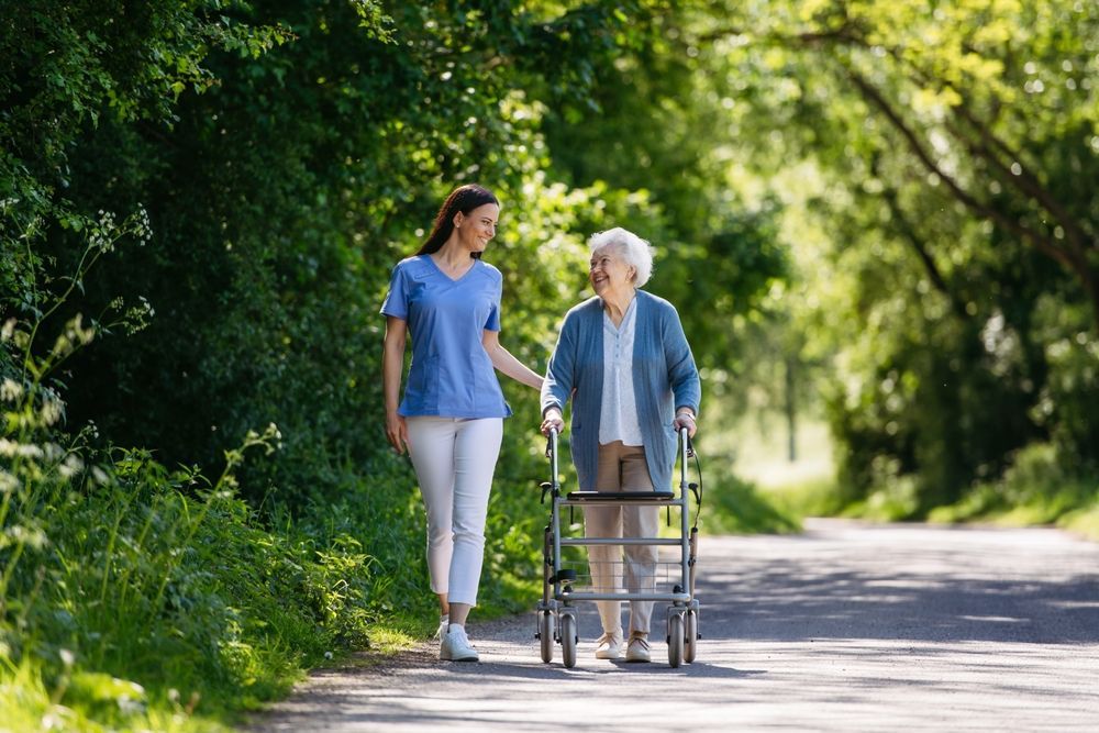 Caregiver assists an older person with a walker on a path, surrounded by trees.