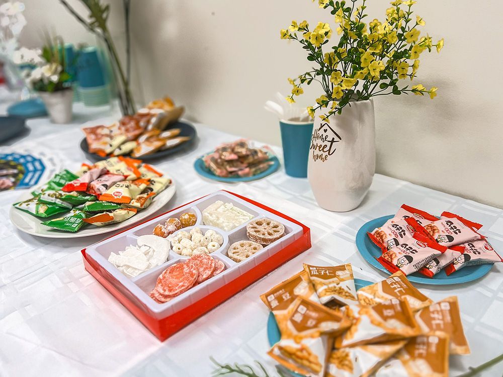 A table with various snacks, including crackers, cookies, and a box of assorted treats, and a vase of yellow flowers.