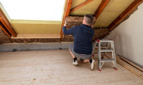 A person wearing a blue hoodie kneels on a wooden attic floor, installing yellow insulation batts between roof rafters.