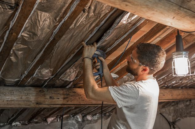 A person wearing protective glasses uses a staple gun to attach foil insulation between exposed wooden attic beams.