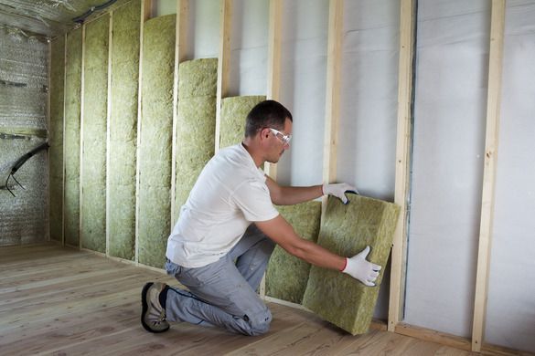 A person wearing safety glasses and gloves installing green insulation panels between wooden wall studs.