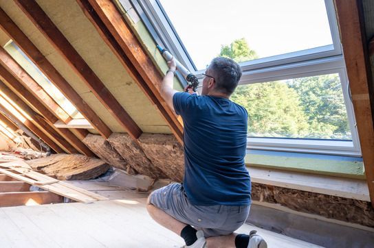 A person kneels in an attic to apply sealant with a caulk gun around the frame of a skylight window.
