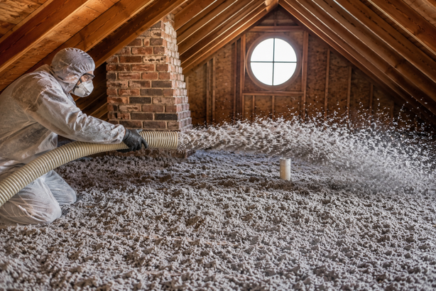 A person in black gloves installing a thick, yellow mineral wool insulation panel onto an attic wall.