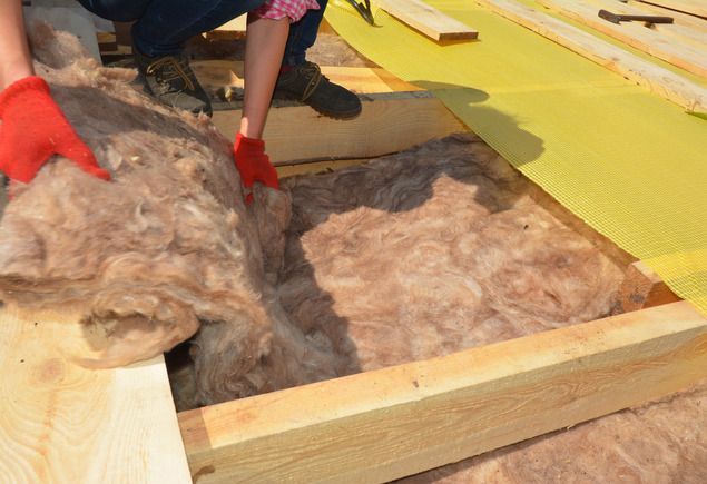 A person wearing red gloves installs pink fiberglass insulation between wooden floor joists.