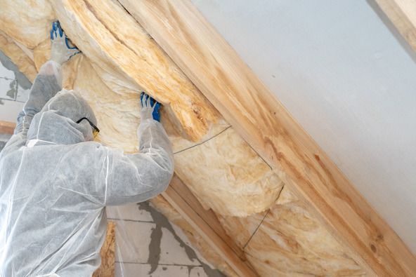 A worker wearing protective gear installs yellow fiberglass insulation between wooden rafters in an attic.