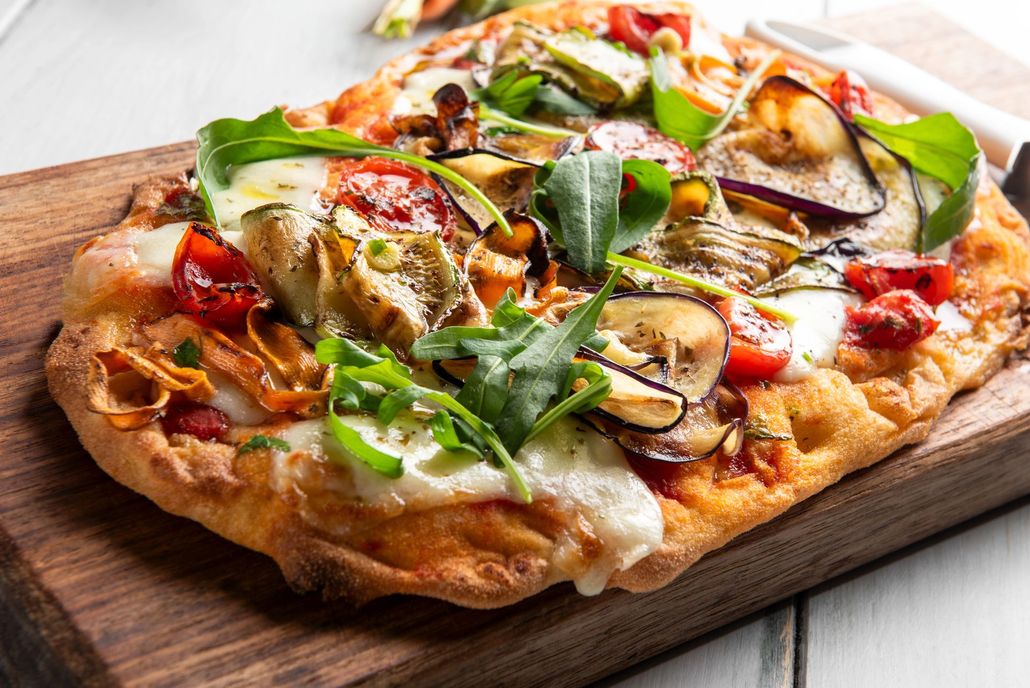 Una pizza su base di focaccia condita con carciofi, verdure arrostite, rucola fresca e formaggio fuso, servita su un tagliere di legno.