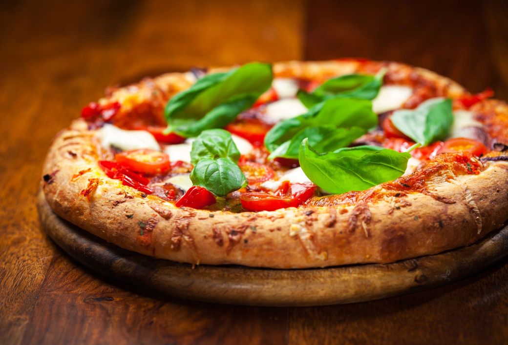 Una pizza rotonda condita con pomodorini rossi, mozzarella bianca e foglie di basilico fresco, adagiata su un tagliere di legno.
