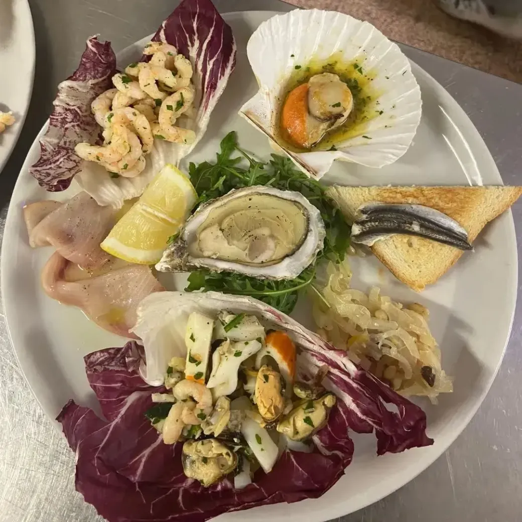 Un piatto con vari frutti di mare, tra cui un'ostrica, una capasanta, gamberi, filetti di pesce e un'acciuga su pane tostato.