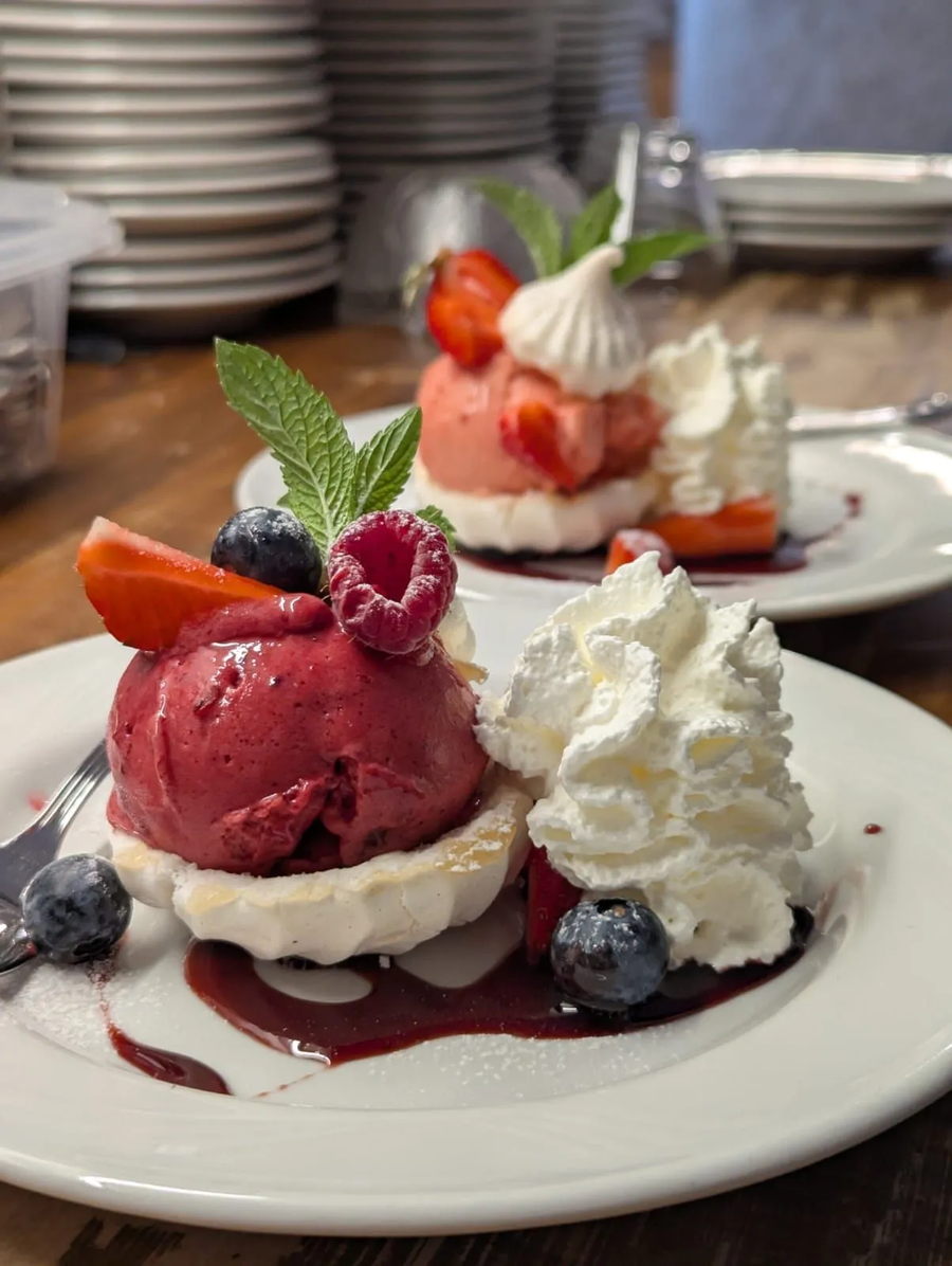 Due piatti con mini pavlova guarnite con sorbetto ai frutti di bosco, panna montata, frutti di bosco freschi e menta, disposti su un tavolo di legno.
