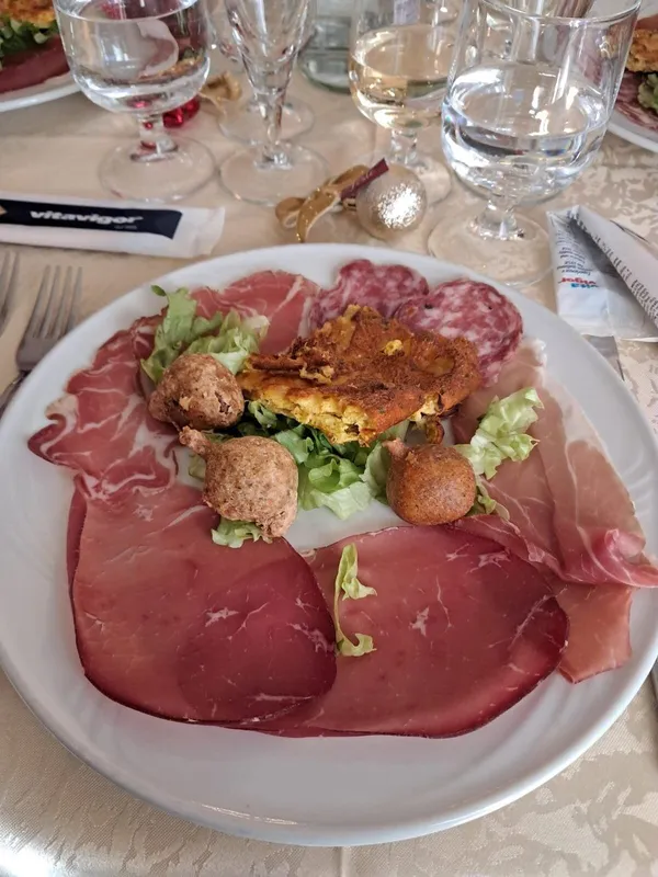 Un piatto bianco colmo di salumi assortiti, tra cui manzo stagionato e salame, con piccole frittelle e guarnizione di lattuga.