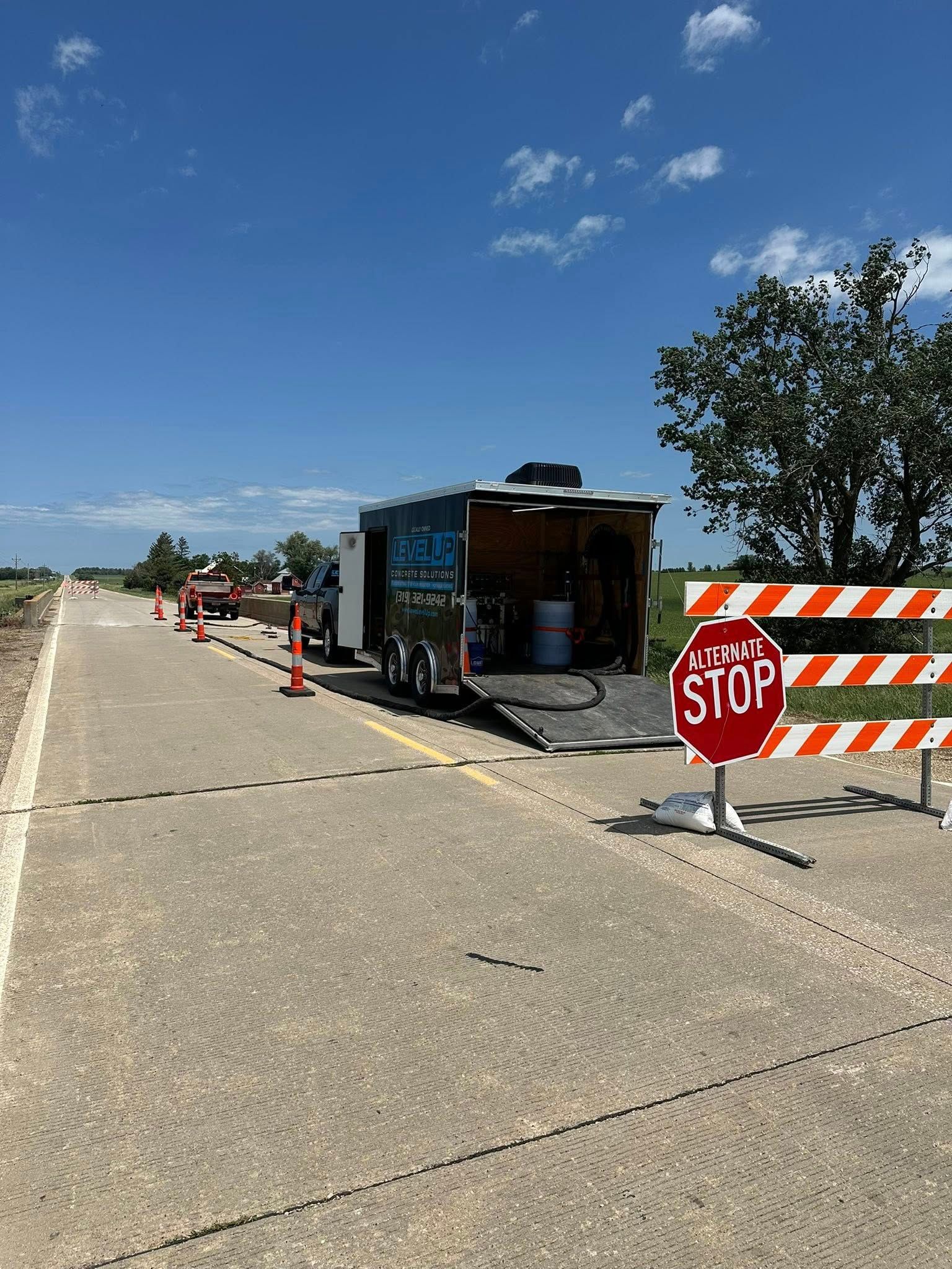 Road work site with trailer, stop sign, cones, and barricade on a concrete road under a blue sky.