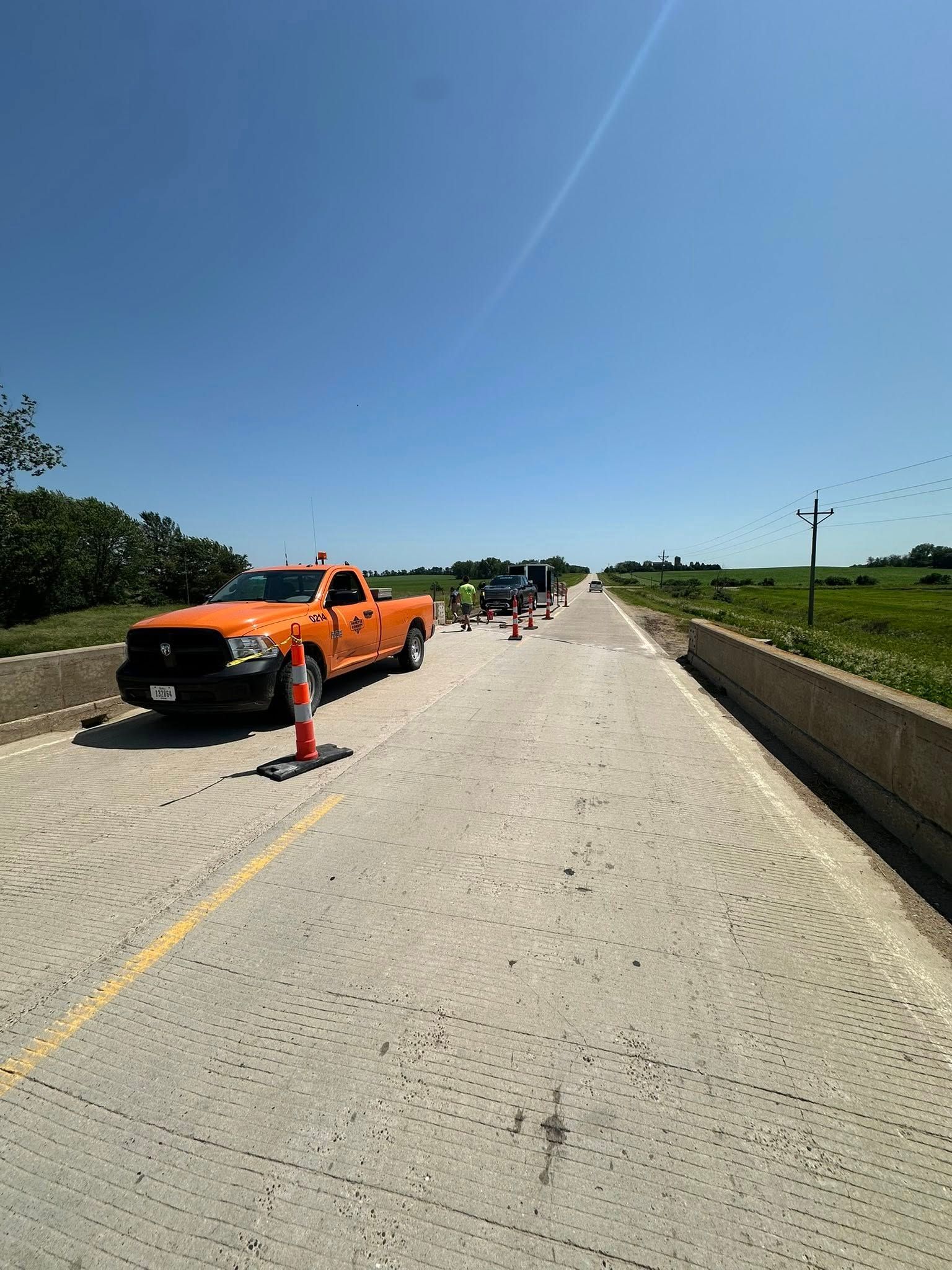 Orange work truck on a bridge, cones blocking a lane. Workers and other vehicles are visible in the distance.
