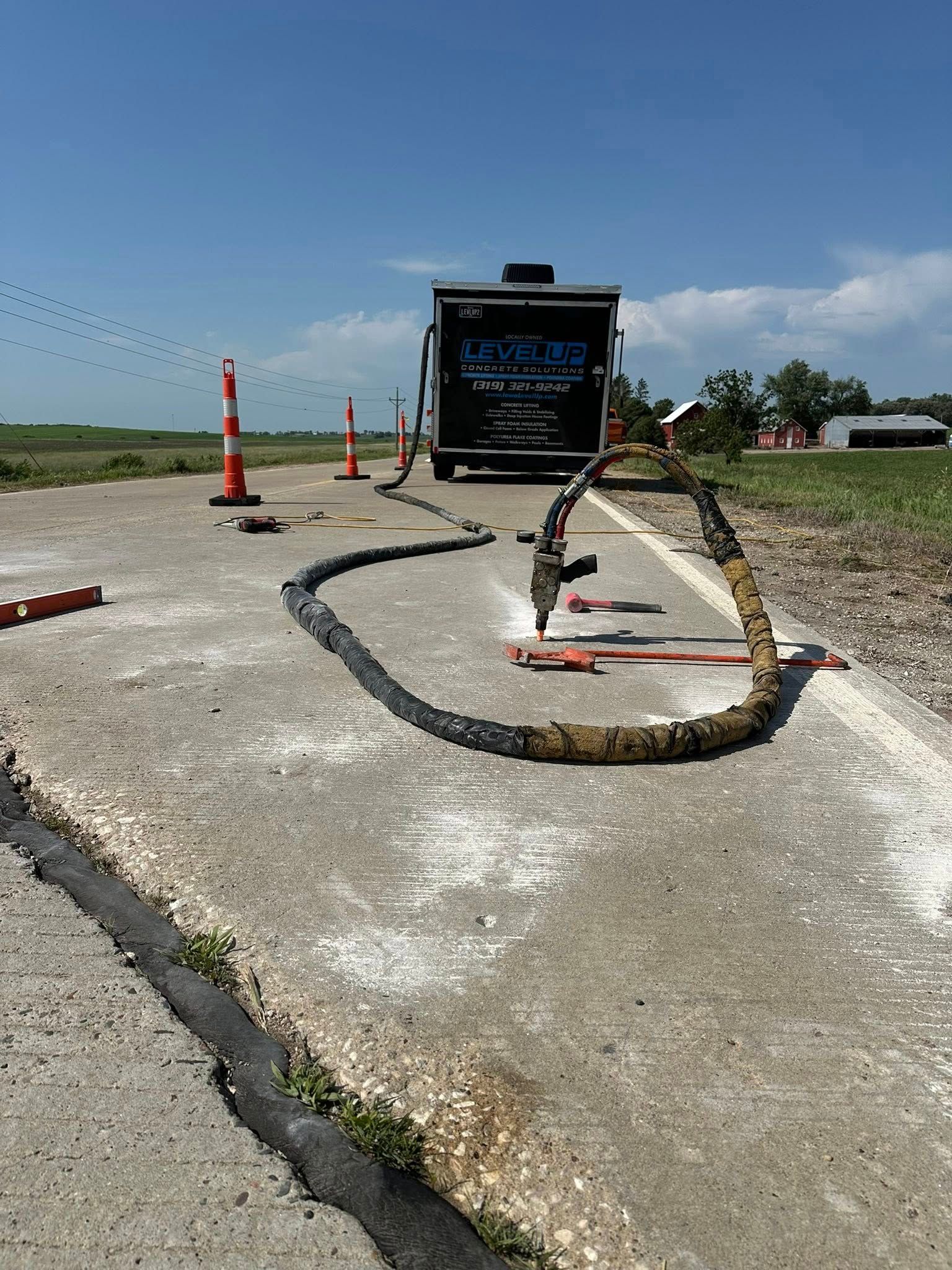 Equipment sealing road cracks. Black hose connected to a trailer, orange safety cones on cracked pavement. Bright sunny day.
