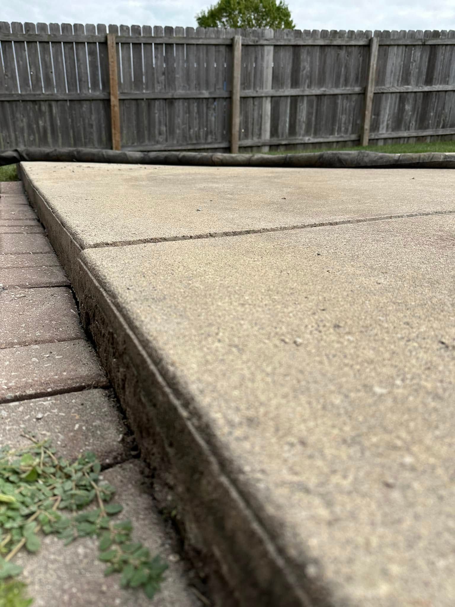 Concrete patio edge with cracks, next to brick walkway and wooden fence.