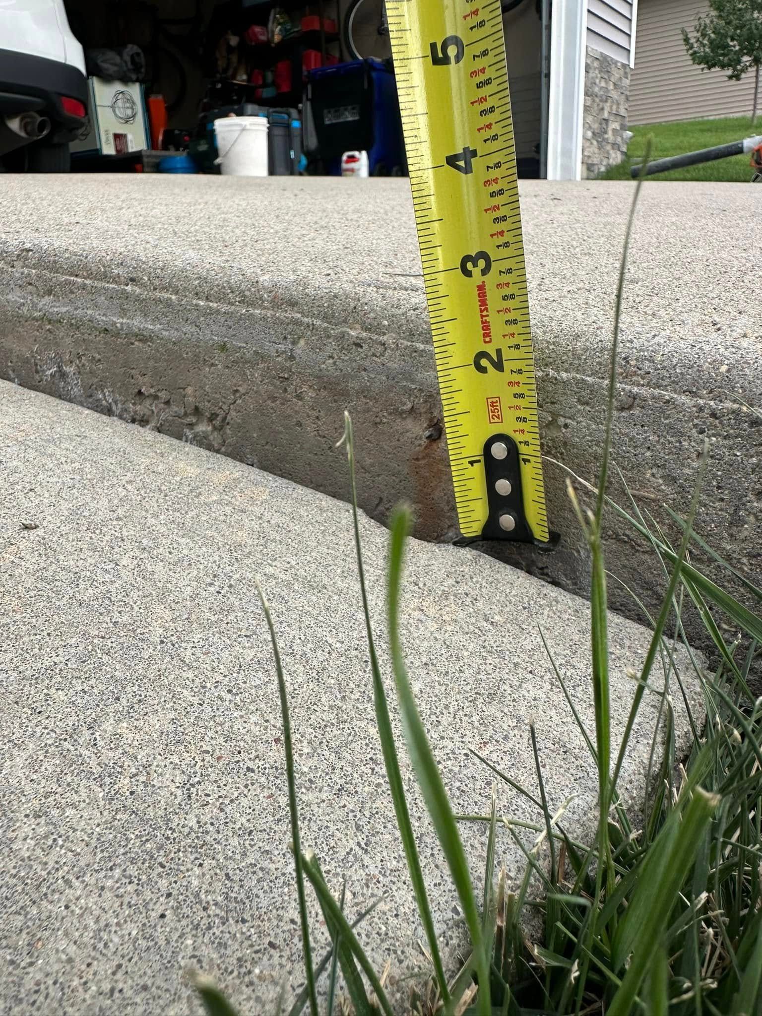 Yellow tape measure showing a 2-inch height difference on a concrete curb with grass.