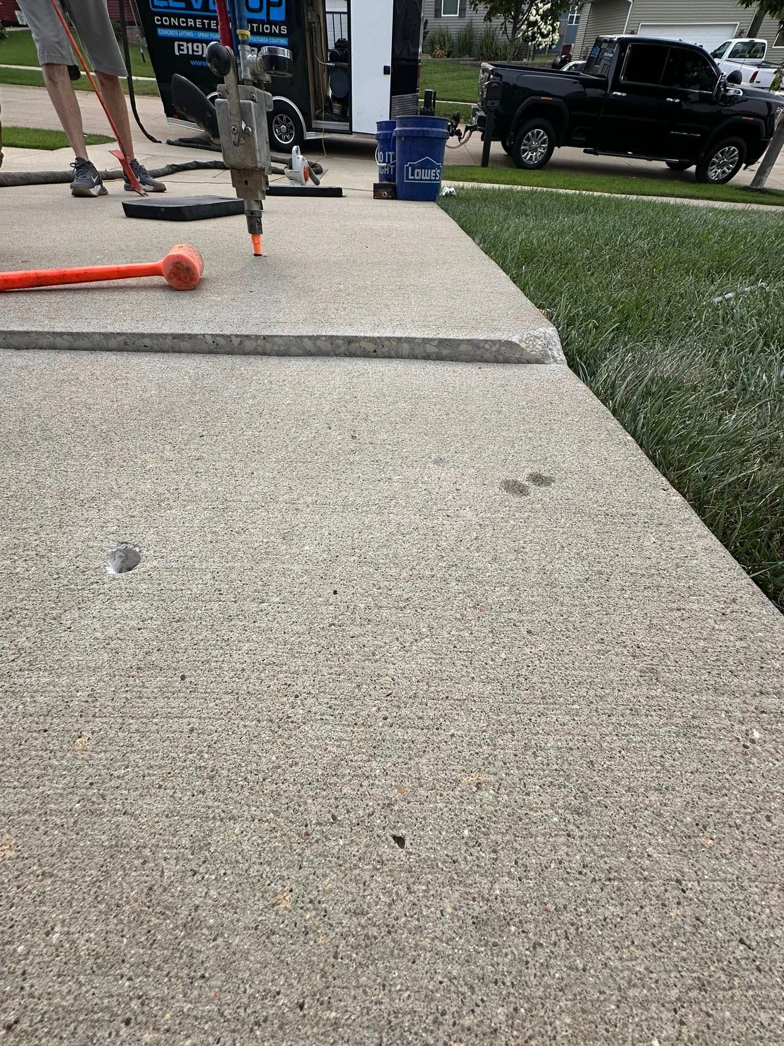 Sidewalk with a raised edge, likely requiring repair; tools and a truck are in the background.