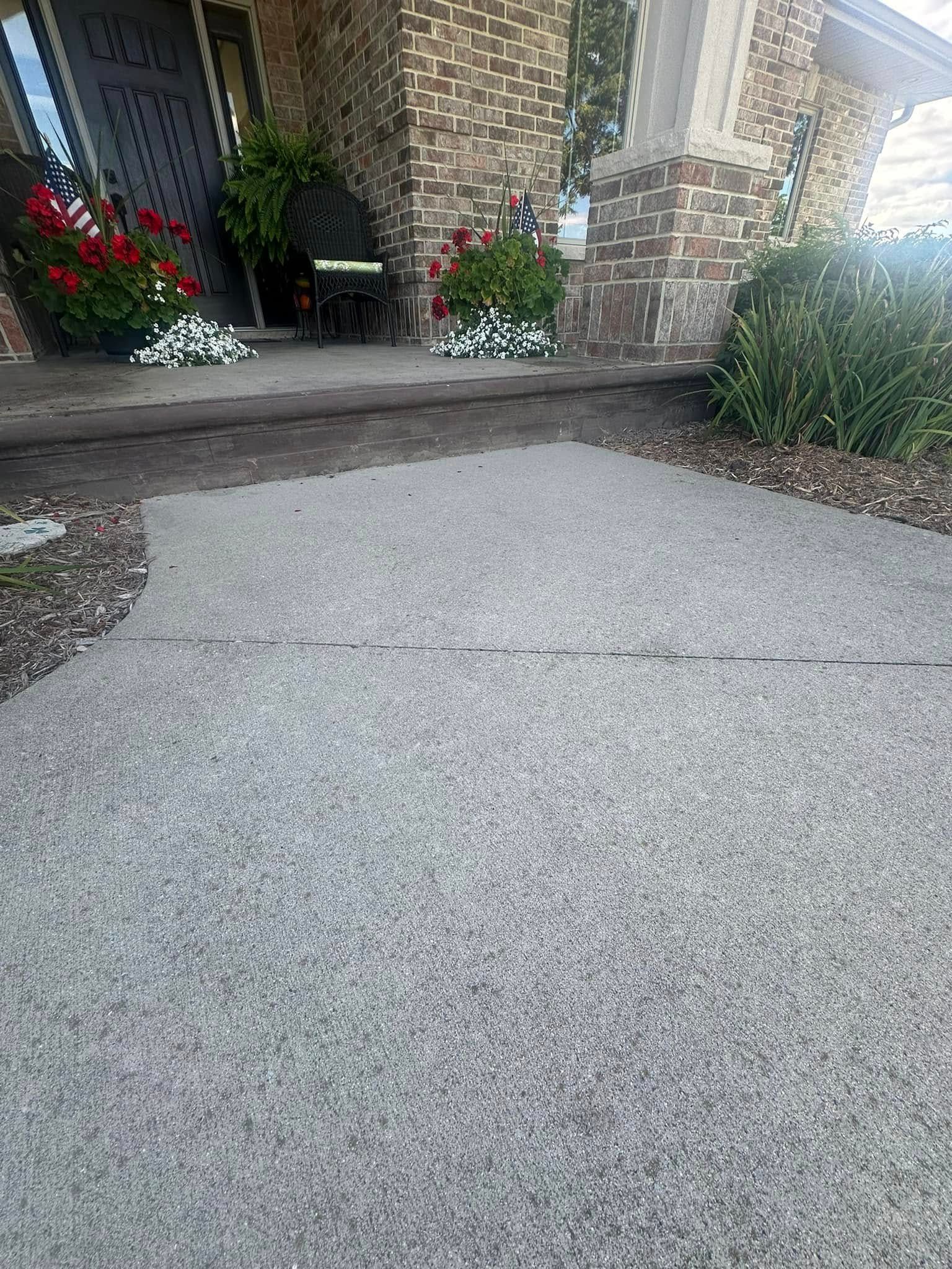 Concrete walkway leading to a house with red brick and flowers.