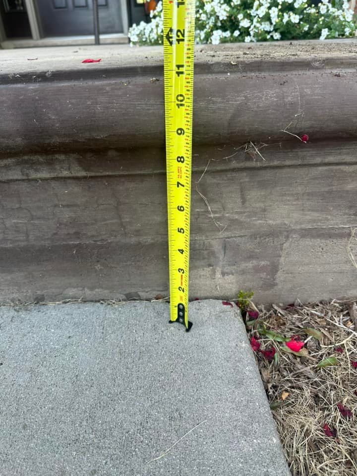 Tape measure showing a step measures 2 inches high. Concrete sidewalk and brown wall in background.