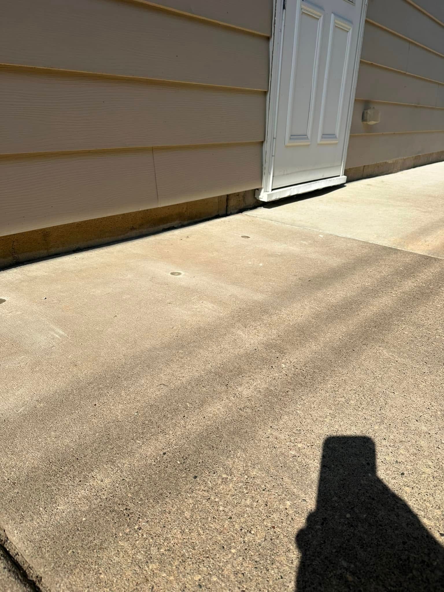 Concrete walkway in front of a tan-colored building with a white door. A shadow is cast across the walkway.