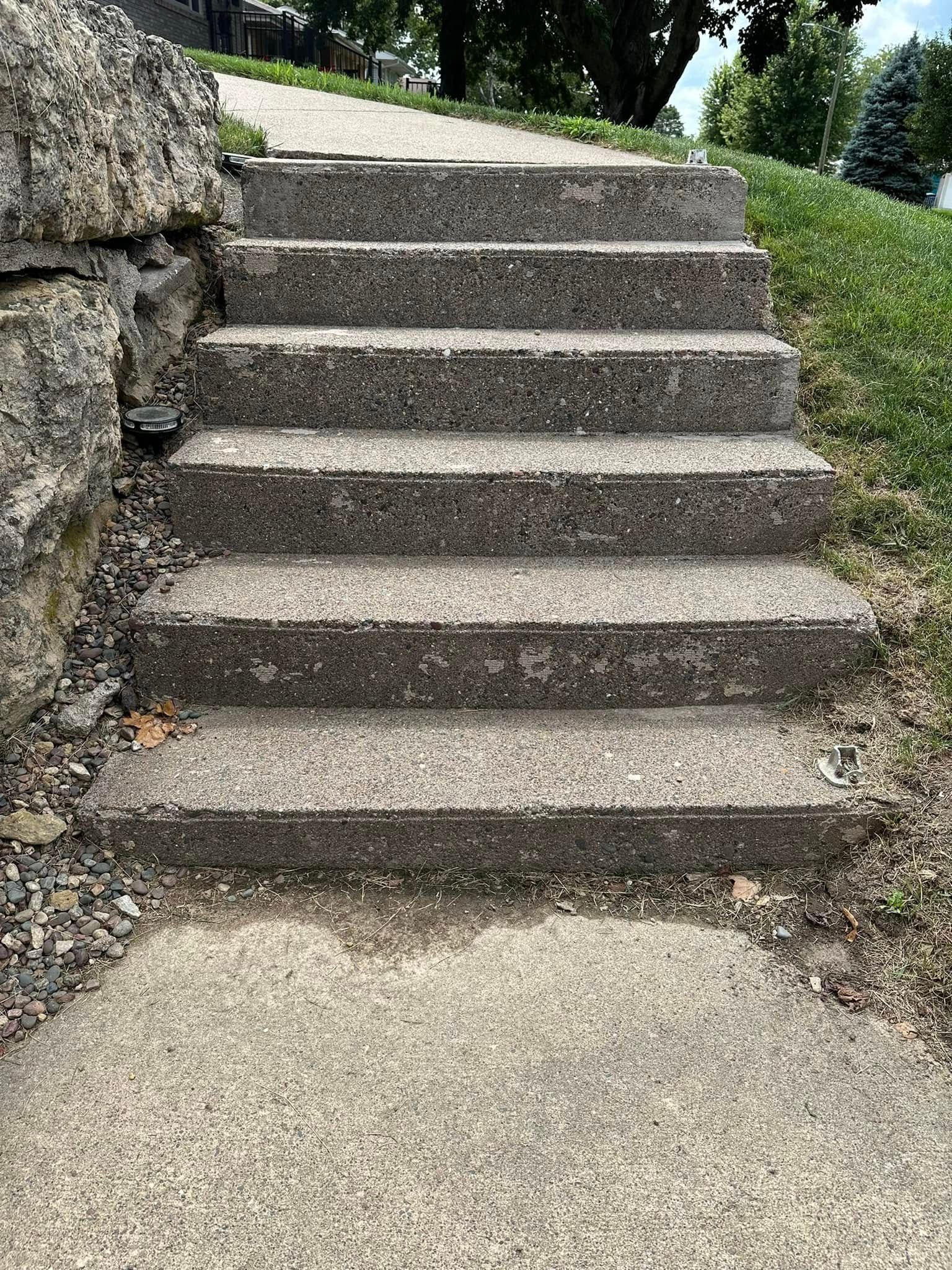 Concrete steps leading uphill from a concrete sidewalk. Stone wall on the left, grass on the right.